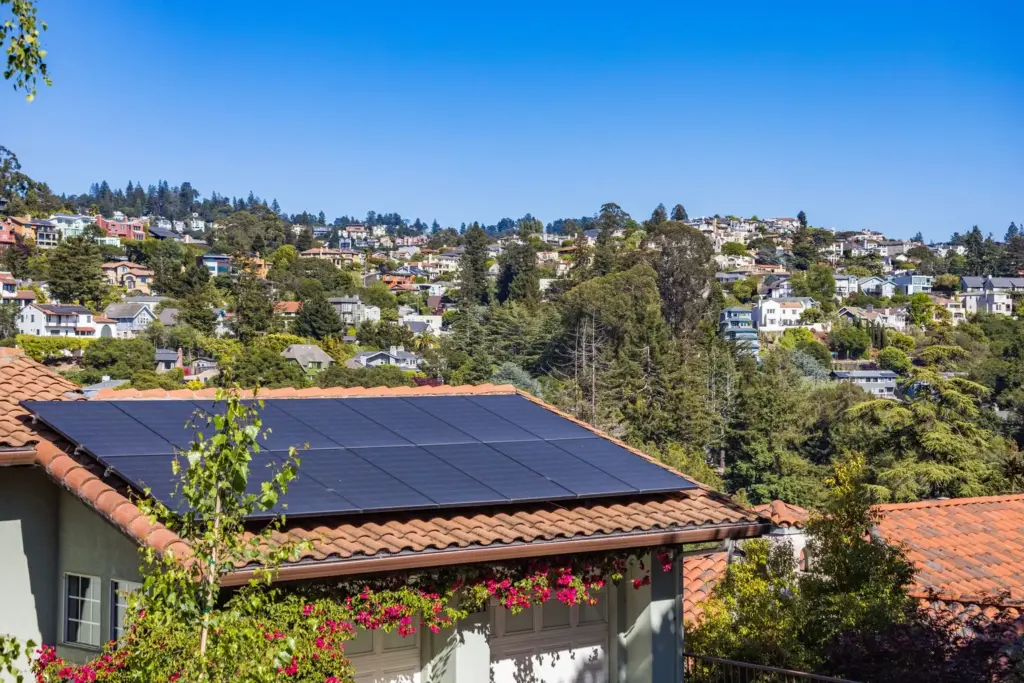 Solar panels installed on a residential roof with a hillside neighborhood and blue sky in the background